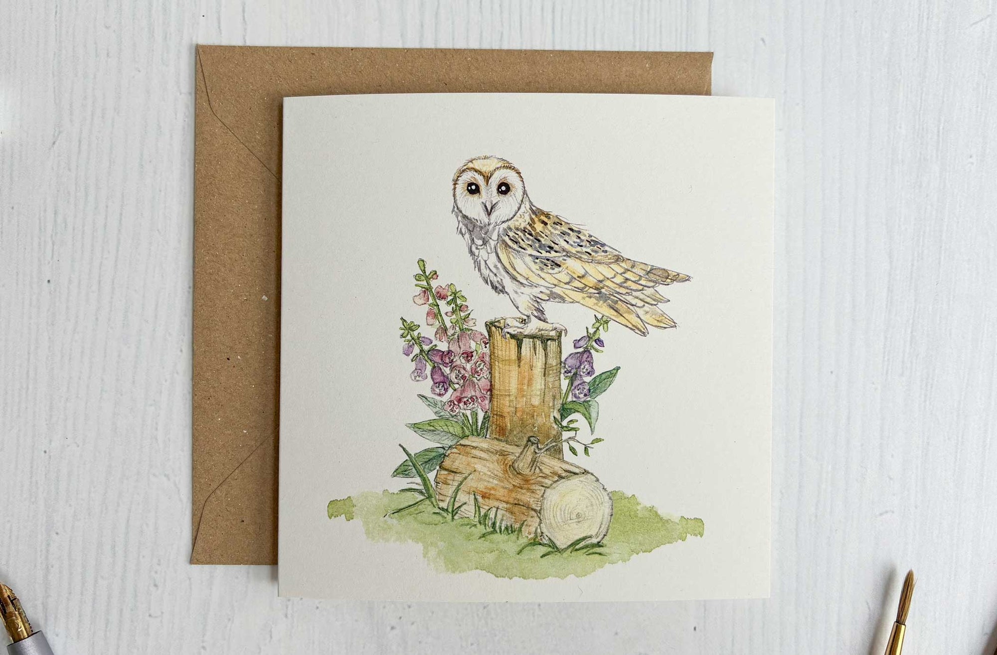 Greeting card with Irish barn owl illustration on a wooden stump, surrounded by flowers and leaves, with a brown envelope and stationery items.