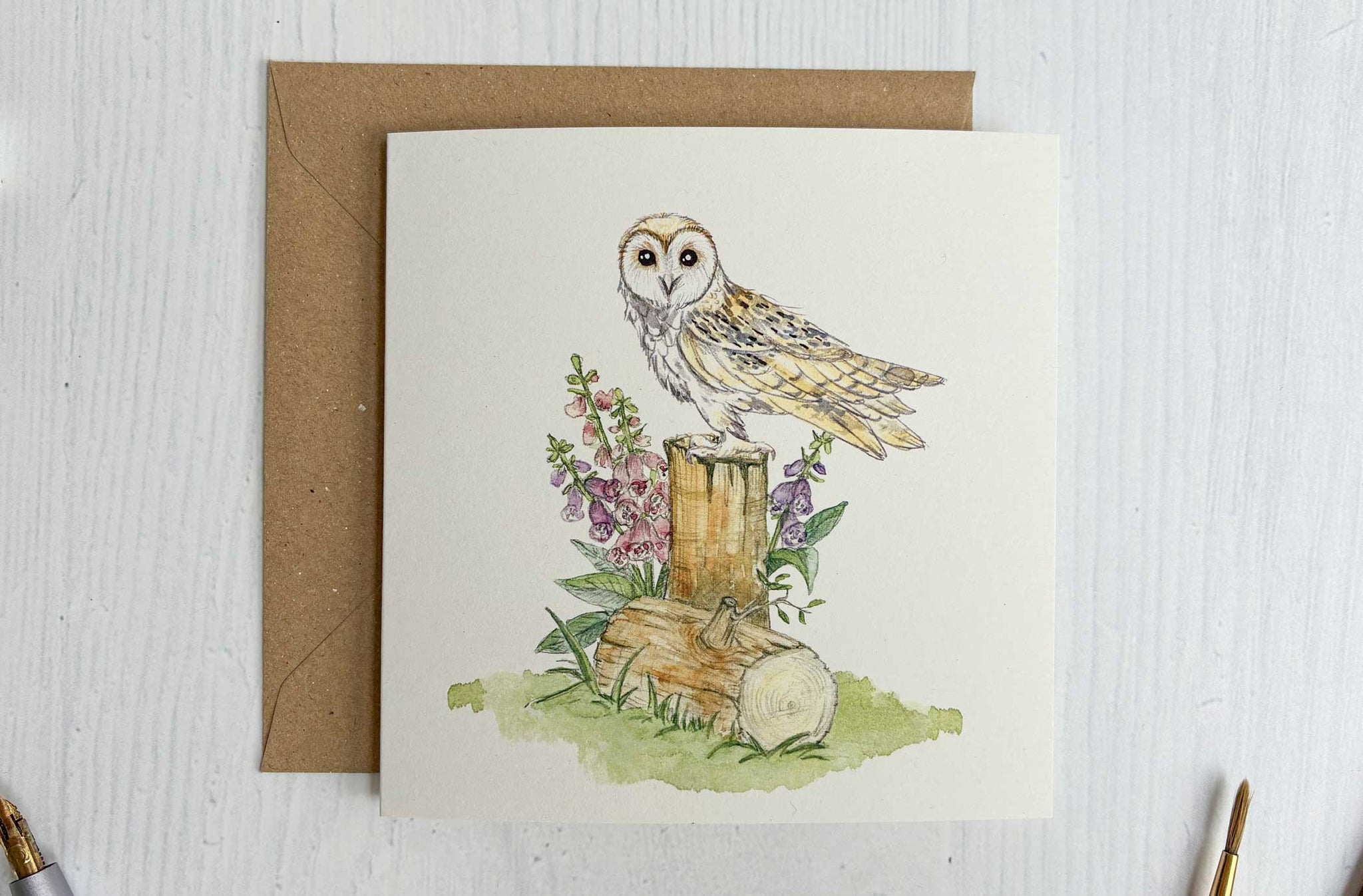 Greeting card with Irish barn owl illustration on a wooden stump, surrounded by flowers and leaves, with a brown envelope and stationery items.