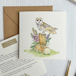 Greeting card with Irish barn owl illustration on a wooden stump, surrounded by flowers and leaves, with a brown envelope and stationery items.