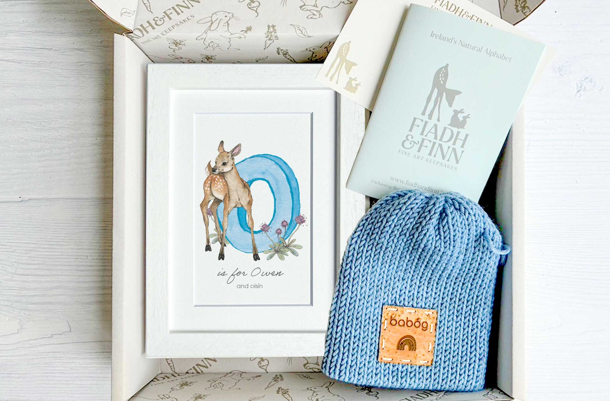 Gift set baby products including a framed print, knitted newborn hat, in box on a wooden surface.