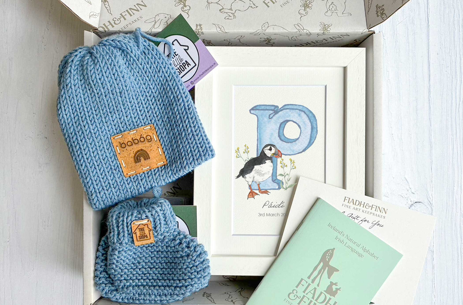 Blue baby hat and booties set with a framed picture of a puffin and book in a box.