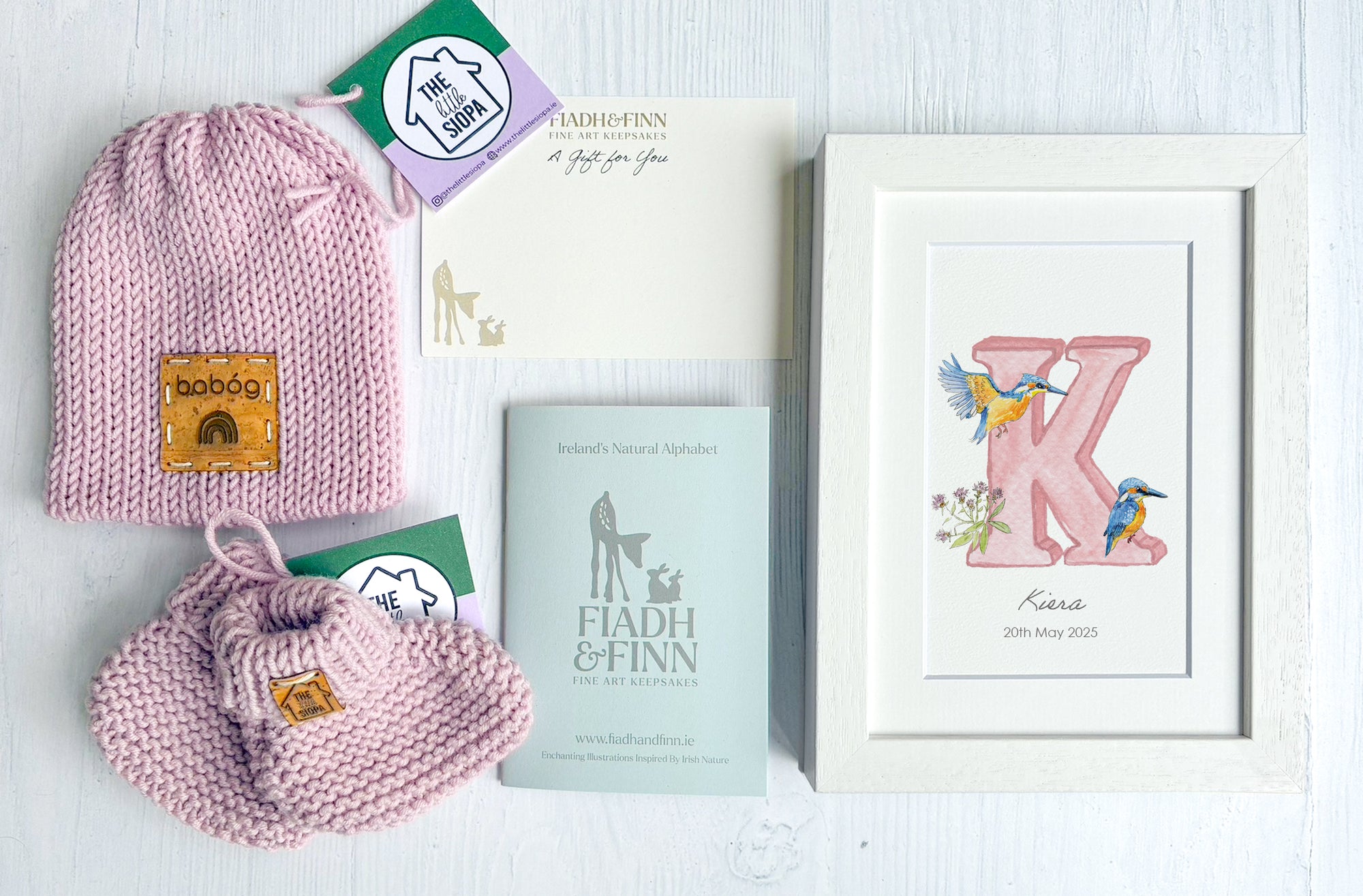 Pink knitted baby hat and booties with branded labels, framed artwork, and books on a light surface.