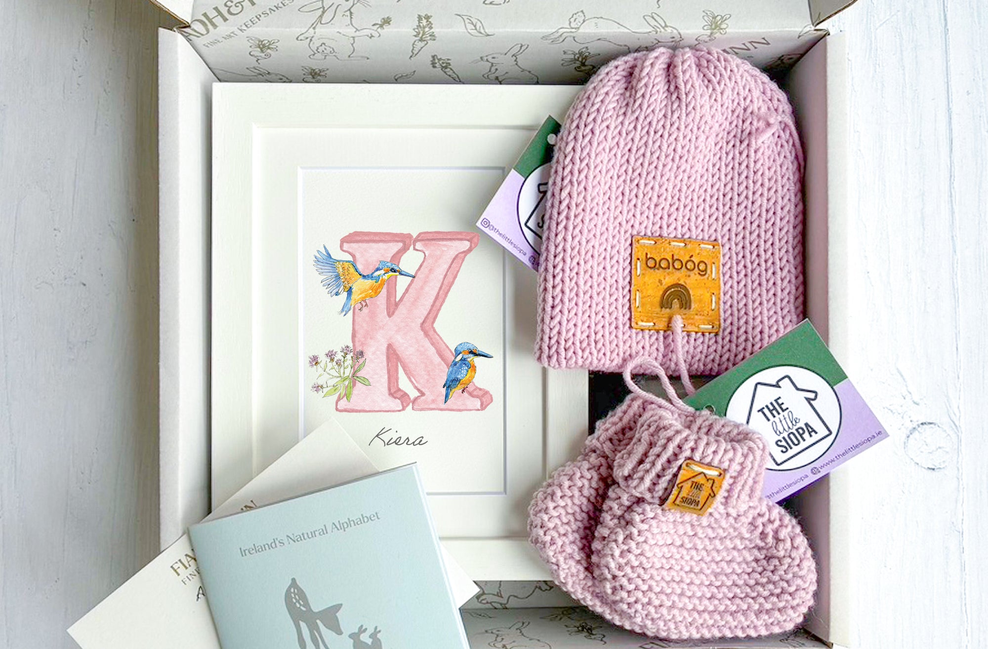 Pink knitted baby hat and mittens set with brand labels in a box.