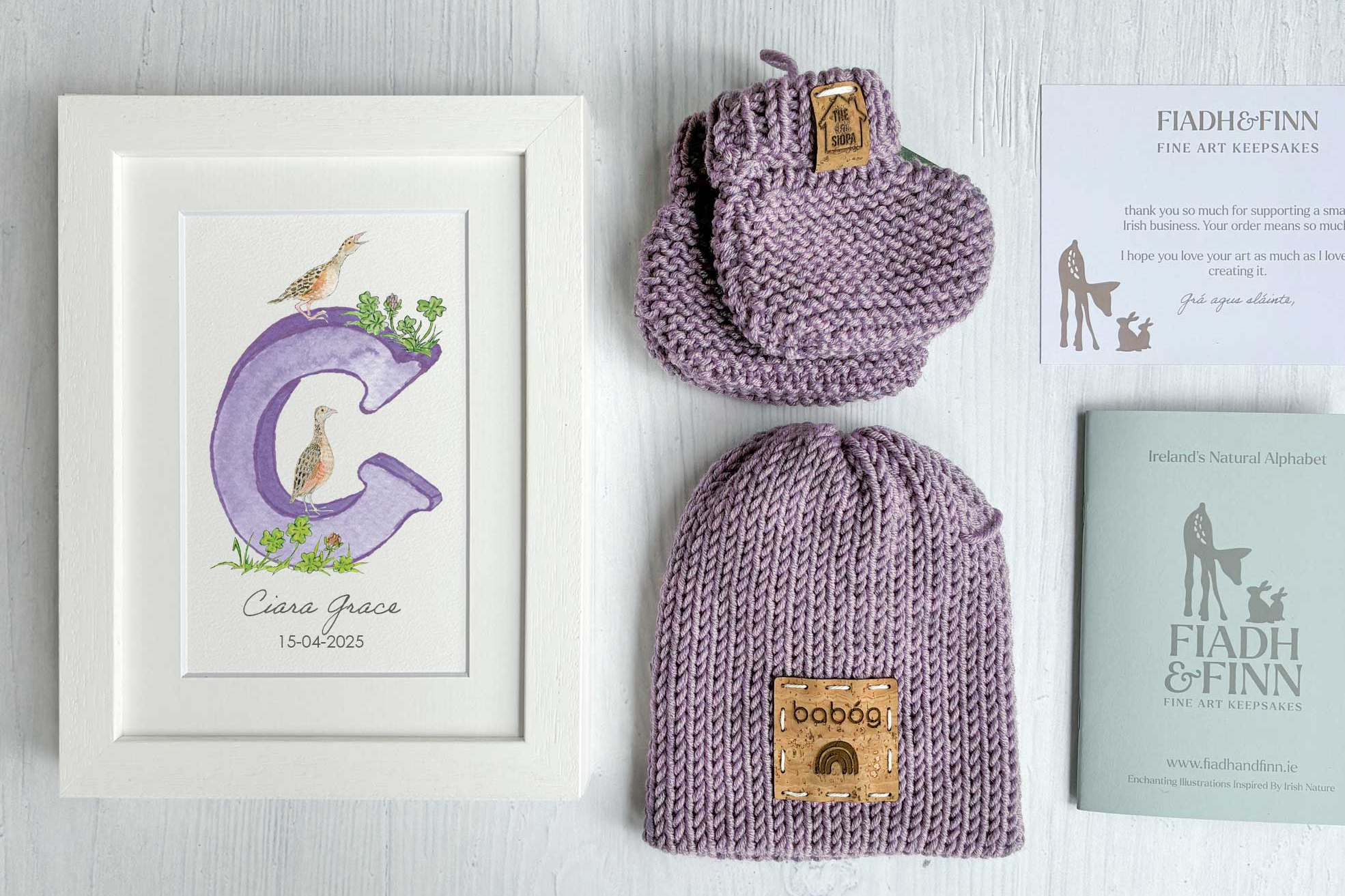 Purple woollen baby hat and baby booties set with a framed print and book on a light gray background
