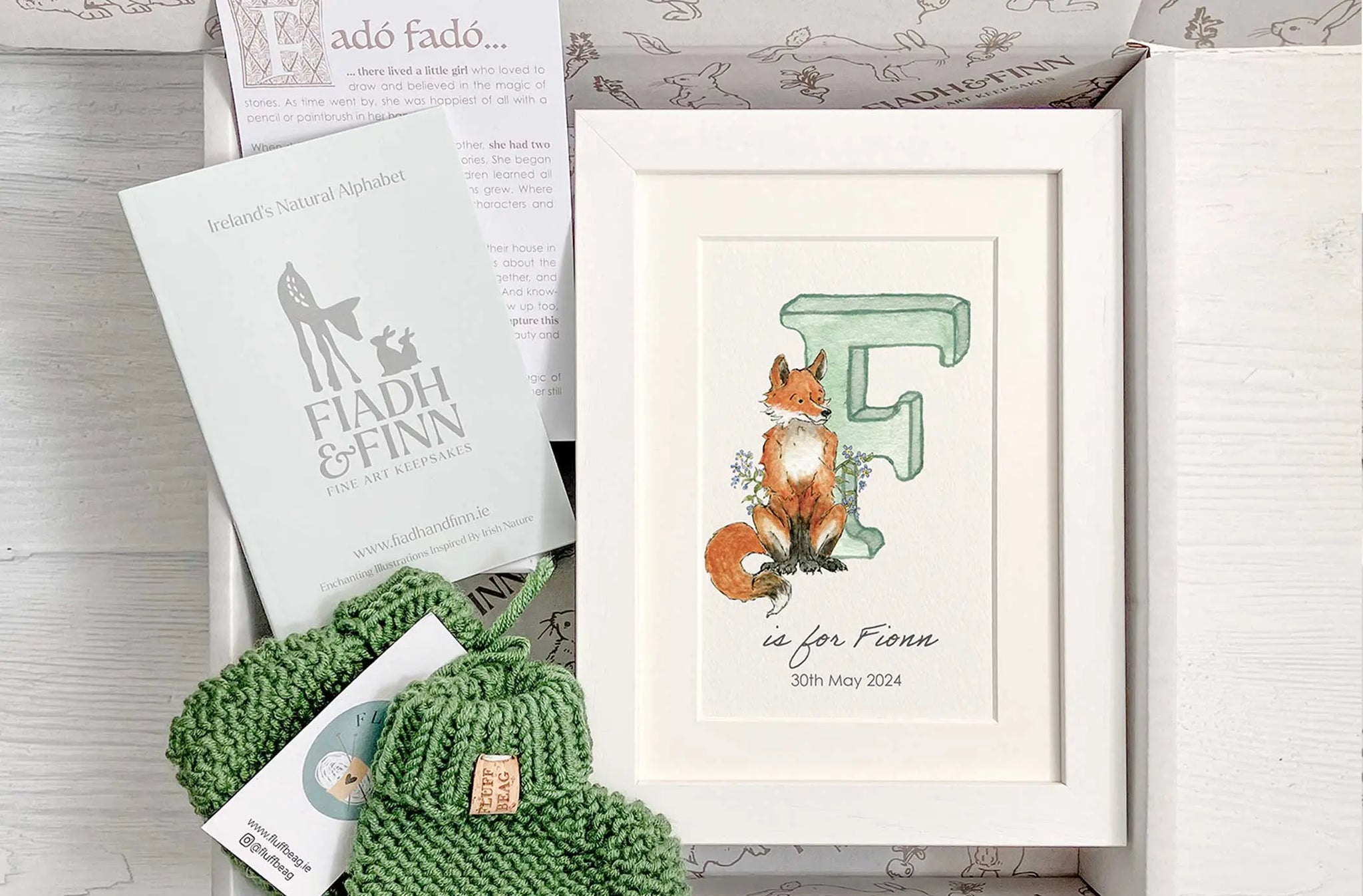 Baby gift box with green watercolour alphabet letter in white frame and green knitted baby booties. Booklet included