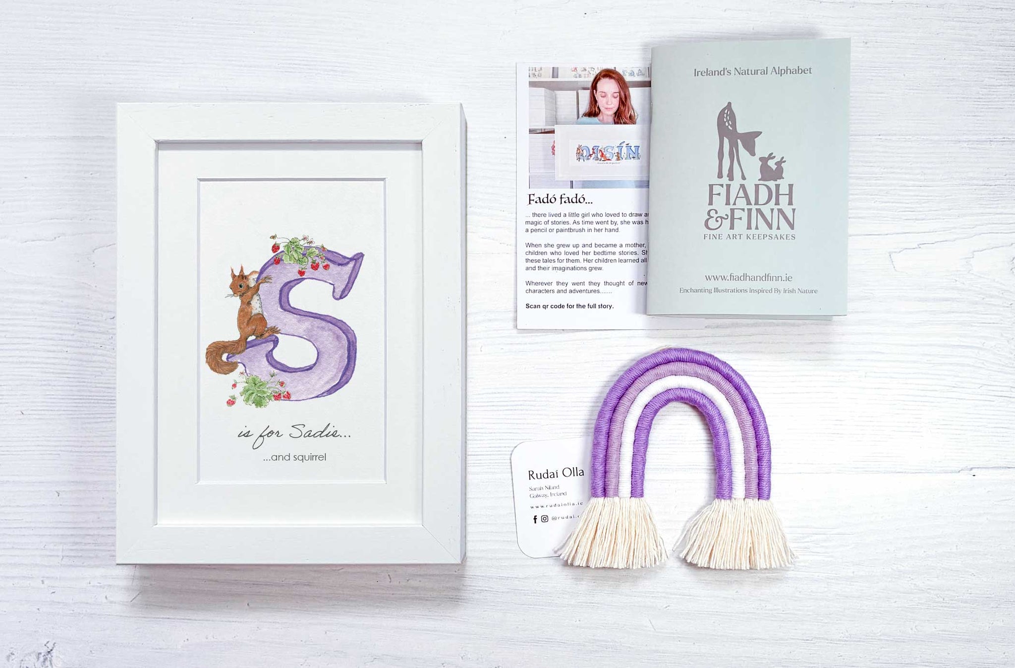 A flat-lay presentation of a personalized baby gift set on a white wood background, including a white framed watercolor letter 'S' print, a purple and white knitted arch with tassels, a small white card, and a grey booklet.