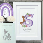 A personalized gift box set featuring a white framed print with a purple letter 'S' and a brown squirrel illustration, a purple and cream tassel rainbow ornament, a mint green 'FIADH & FINN' booklet, and printed cards inside a light wood box.
