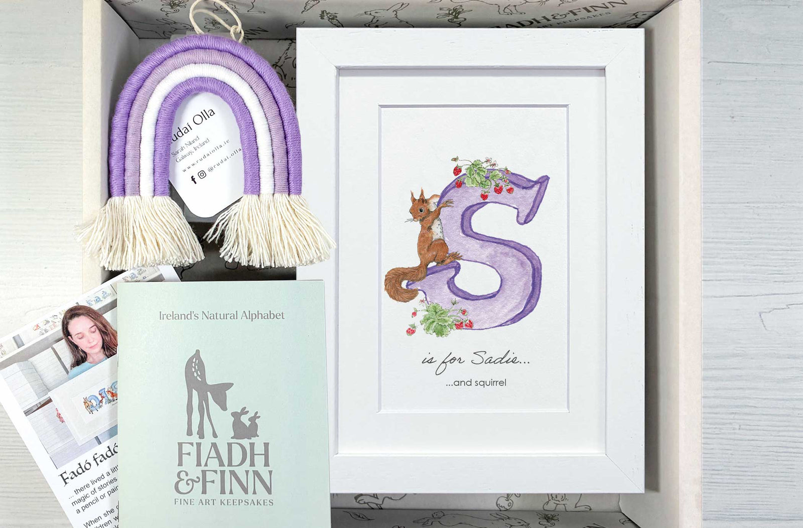 A personalized gift box set featuring a white framed print with a purple letter 'S' and a brown squirrel illustration, a purple and cream tassel rainbow ornament, a mint green 'FIADH & FINN' booklet, and printed cards inside a light wood box.