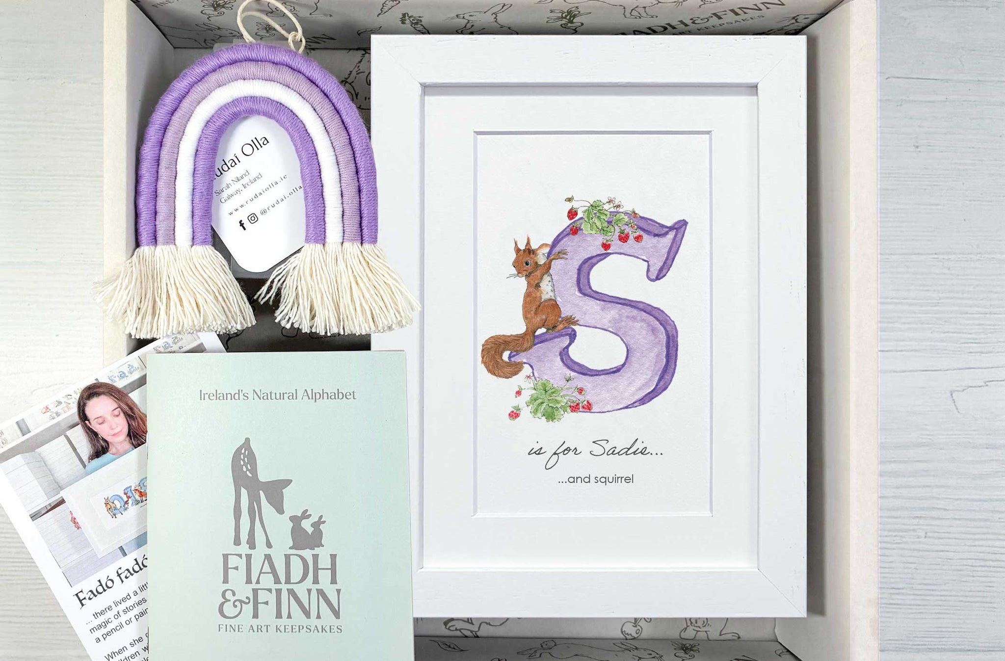 A personalized gift box set featuring a white framed print with a purple letter 'S' and a brown squirrel illustration, a purple and cream tassel rainbow ornament, a mint green 'FIADH & FINN' booklet, and printed cards inside a light wood box.