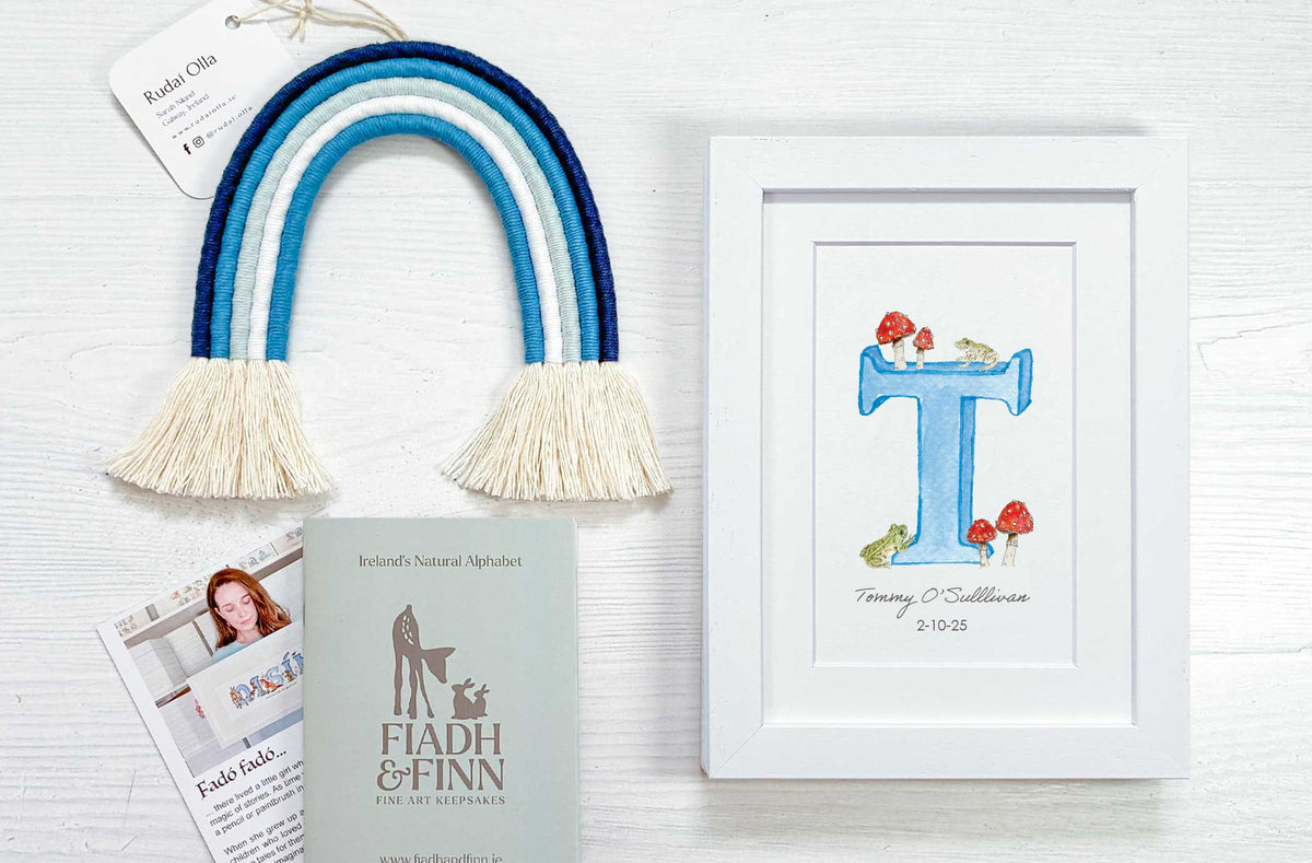 Personalized alphabet art print in a white frame next to a handmade blue and white macrame rainbow with cream tassels, arranged on a white wood background with Fiadh & Finn branding materials.