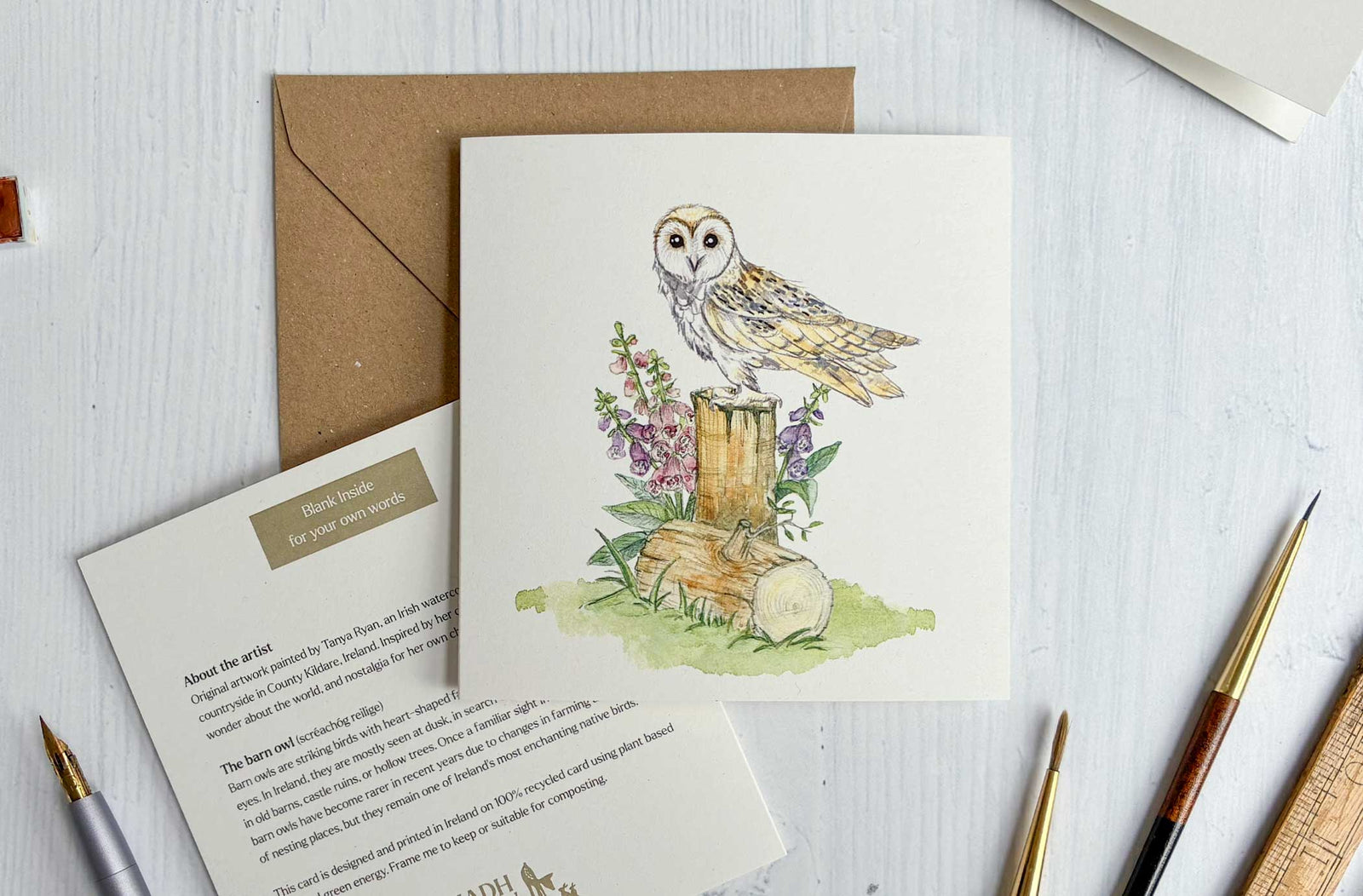 Greeting card with Irish barn owl illustration on a wooden stump, surrounded by flowers and leaves, with a brown envelope and stationery items.
